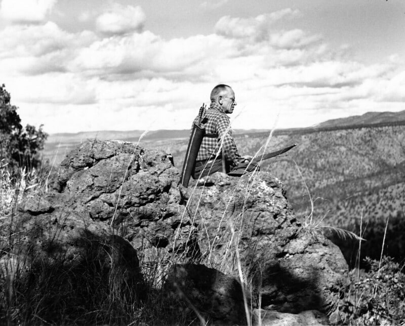 A black and white image of Aldo Leopold, an early proponent of the sustainability movement, sitting on a rock in the mountains.