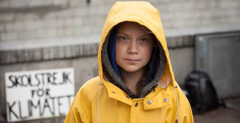 Greta Thunberg standing in a yellow raincoat with her hood on. There is a steamer behind her back saying “School strike for climate” in Swedish